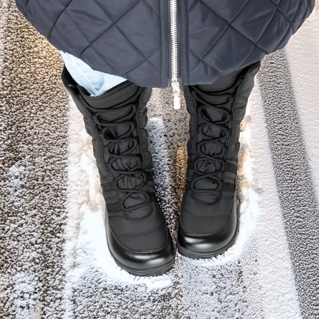 Vaistoa Hanki – korkeavartiset paljasjalkakengät talveen, väri musta. Käyttökuva: ylhäältä päin kuvattuna, henkilö sinisissä farkuissa ja mustassa toppatakissa kävelee lumisella tiellä talvisessa kaupunkiympäristössä.