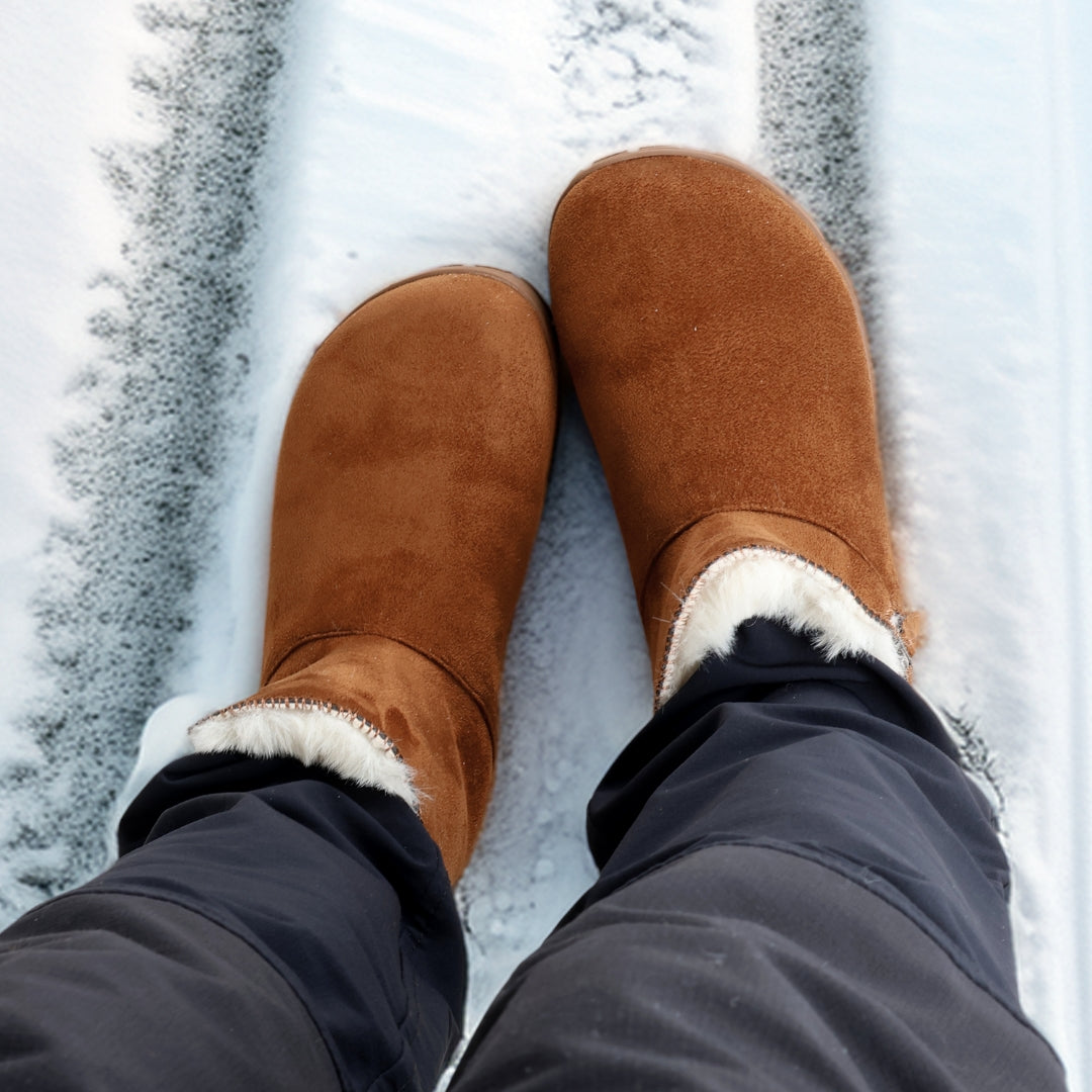 Vaistoa Huurre – paljasjalkabootsit talveen, väri karamellinruskea. Käyttökuva: ylhäältä päin kuvattuna, henkilö seisoo lumella Huurre-bootsit jalassa – karvavuori ja lämmin rakenne näkyvät selkeästi.