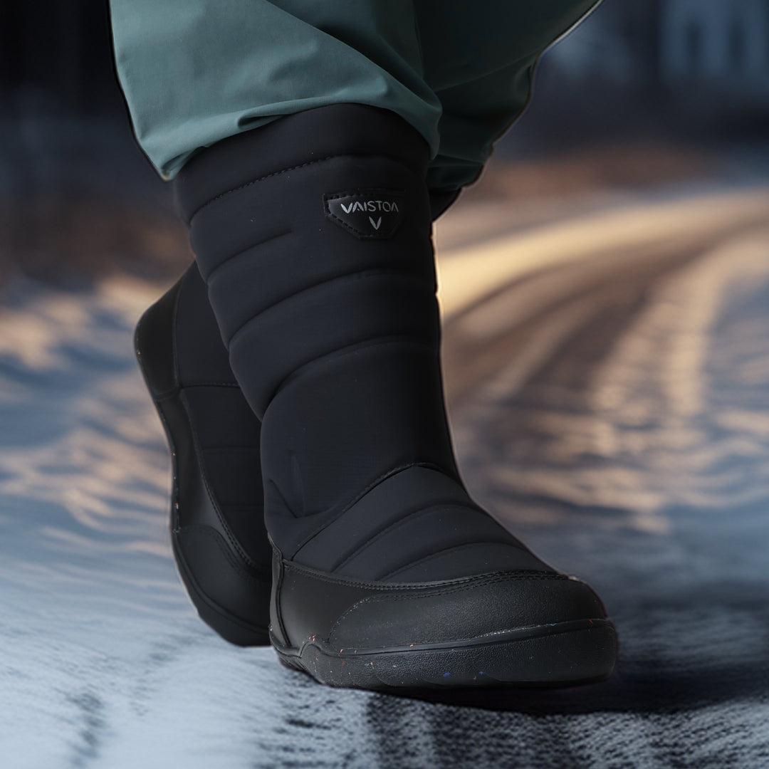 Vaistoa Kinos – lasten korkeavartiset paljasjalkakengät talveen, väri musta. Käyttökuva: etuviistosta kuvattuna sinisten housujen kanssa talviympäristössä.